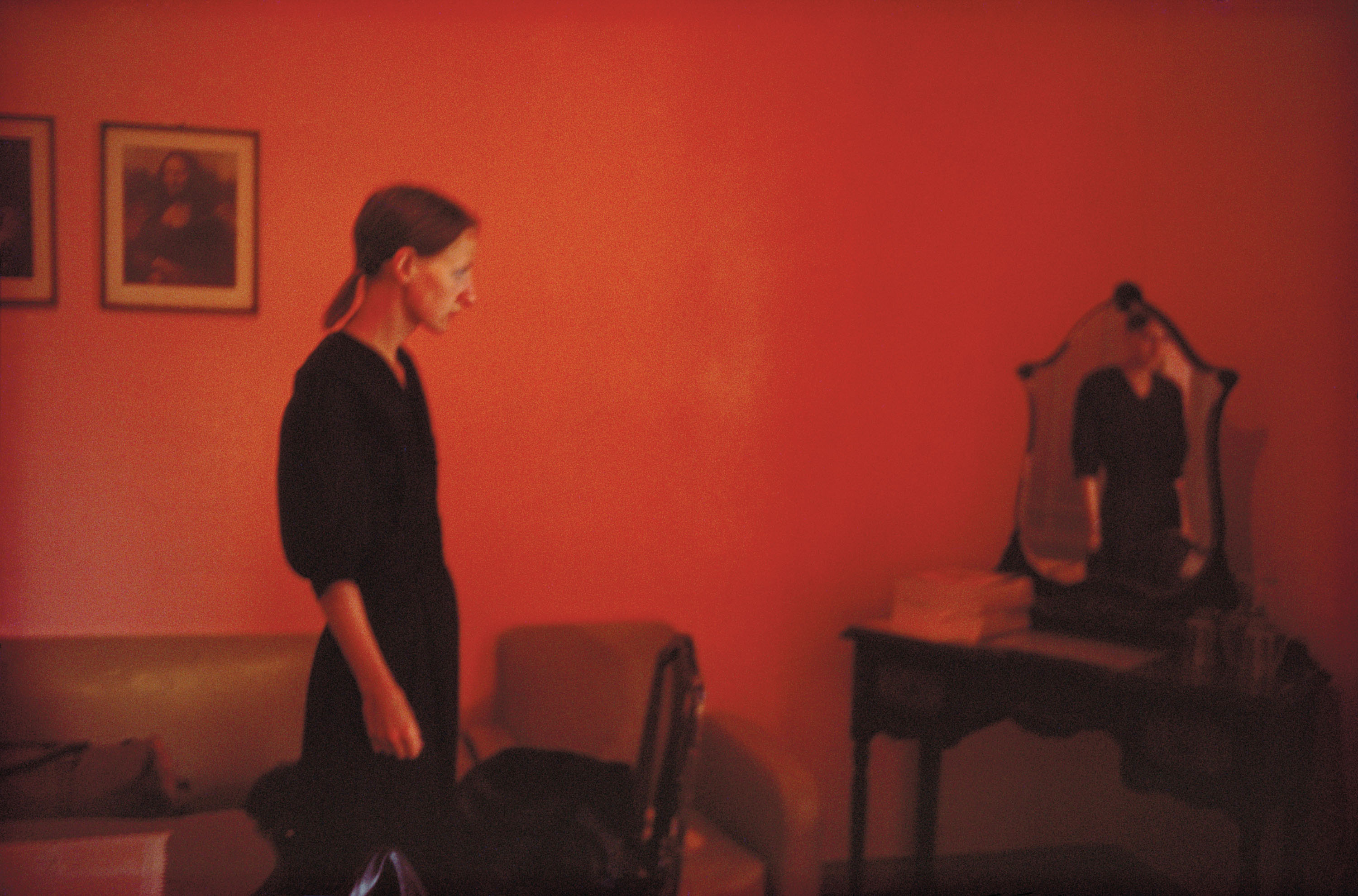 A woman in a black dress stands in profile in a room with deep red walls, her reflection appearing in an ornate vanity mirror across the room.
