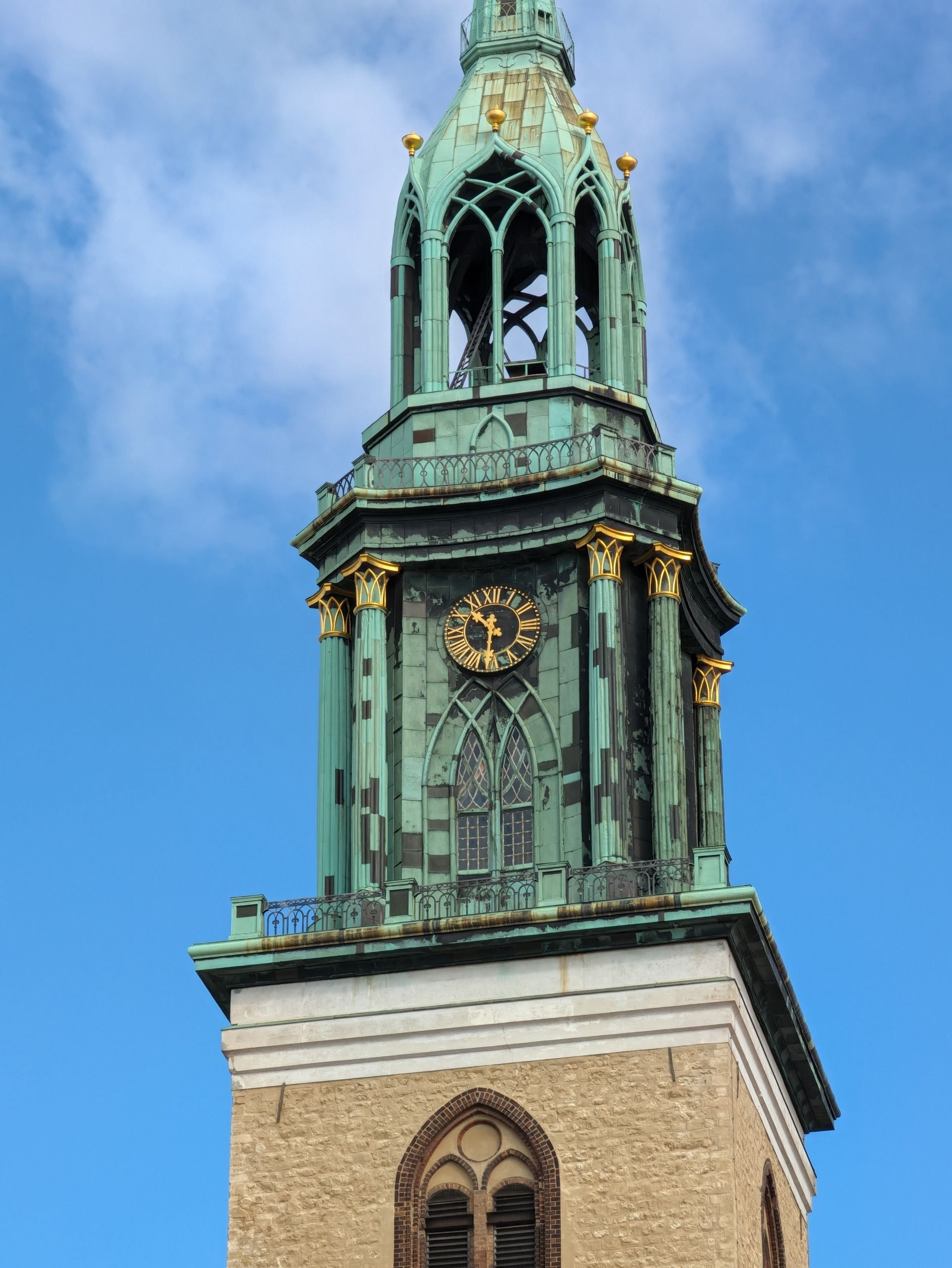 Telephoto view of an ornate green copper church tower and clock against a blue sky.