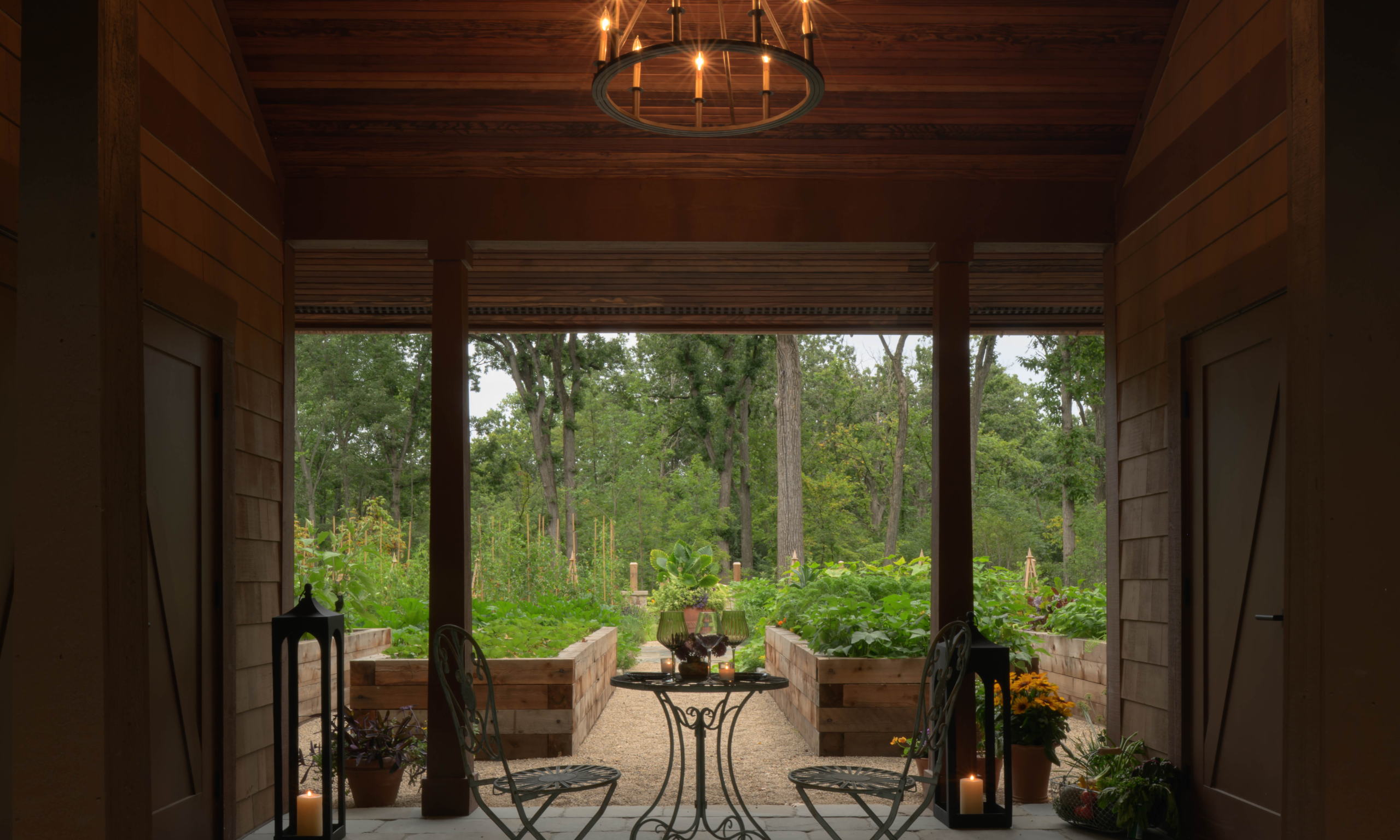Interior of a garden barn, with a chandelier and iron table and chairs, looking onto kitchen garden