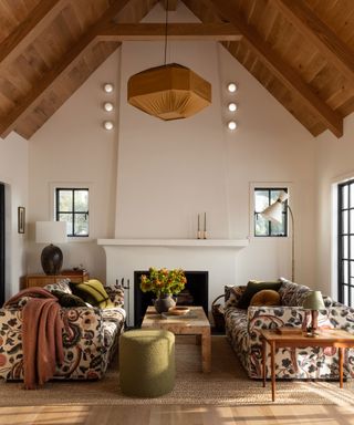 Large living room with wooden vaulted ceiling, white painted walls, black window frames, light wooden flooring, textured rug, floral patterned sofas, marble coffee table, green pouf, fireplace, painted white surround, light with fabric shade in brown colorway