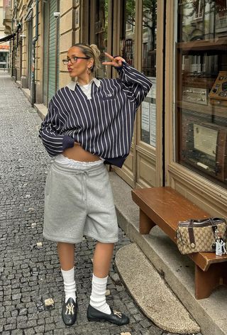 A woman wearing striped rugby polo with gray sweatshorts, white socks, and black loafers.