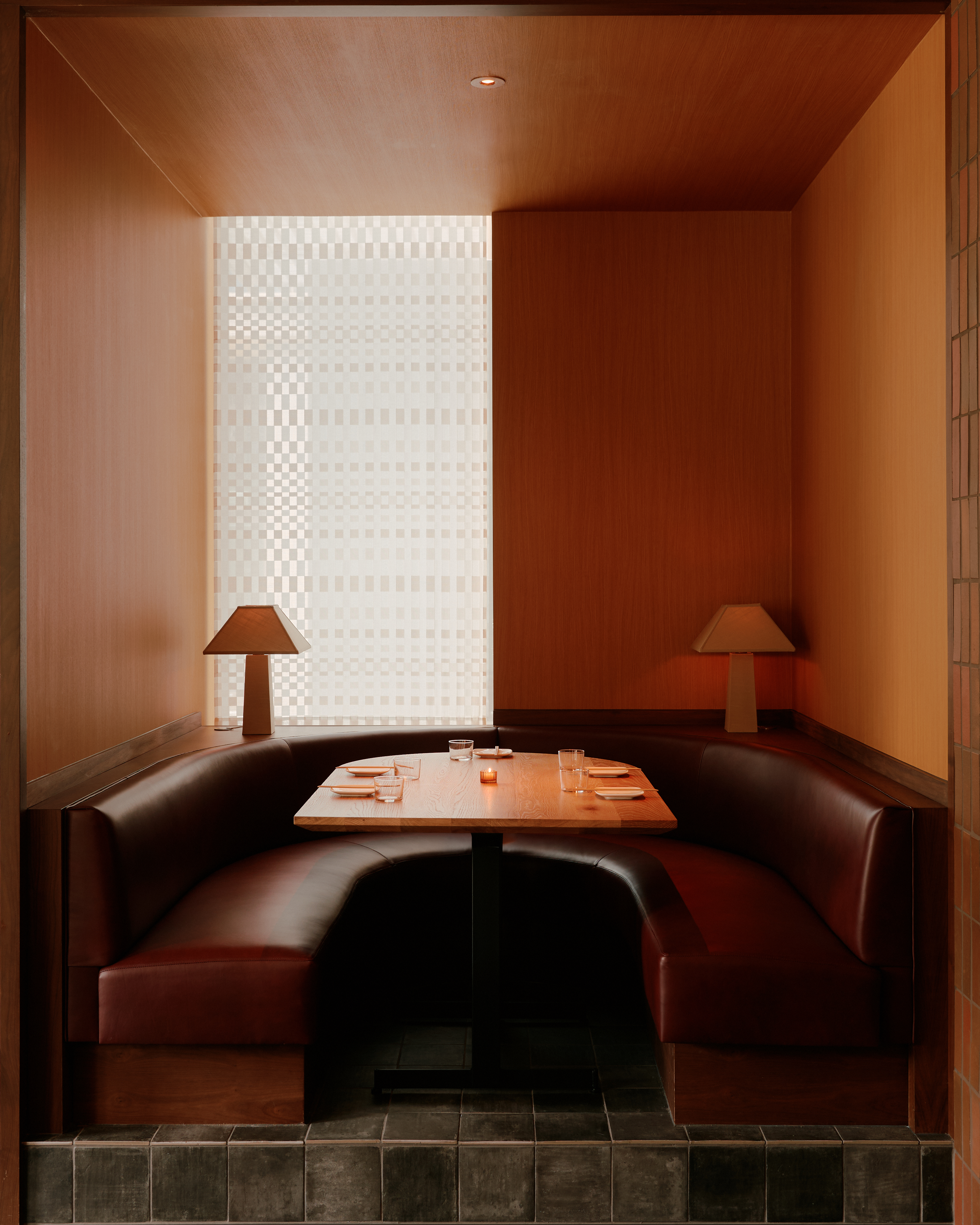 A brown leather banquette seat in a restaurant, decorated with two sculptural table lamps, wooden walls, tiled detailing, and a reversed U-shaped table.