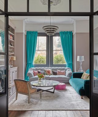 pastel living room ideas, pastel pink living room with turquoise blue drapes and couch, grey couch, grey rug, glass coffee table, built in shelving in alcoves, chandelier, view through crittall doors