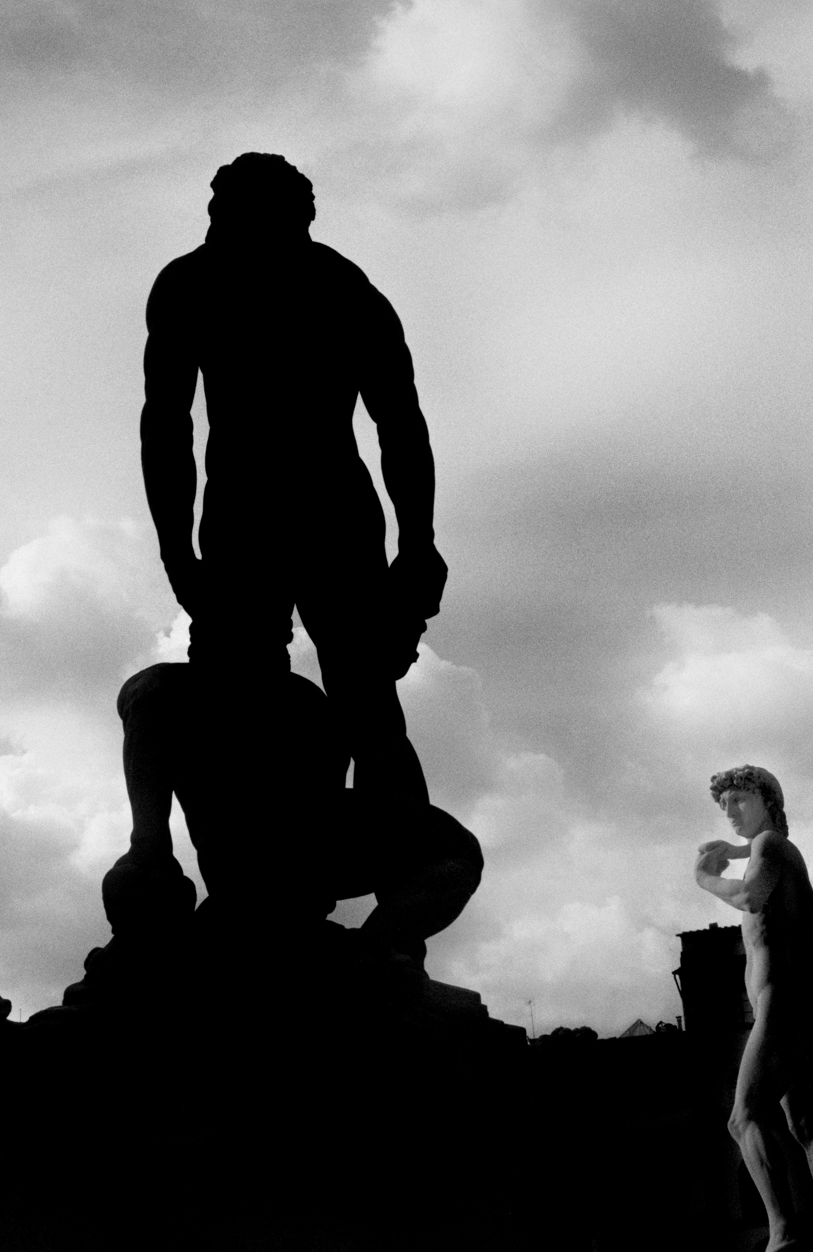 A towering statue of Hercules is silhouetted against a cloudy sky, while a pale statue of David stands at the edge of the frame.