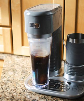 Making iced coffee using the Mr. Coffee 4-in-1 Single-Serve Latte Lux, Iced, and Hot Coffee Maker which is placed on a granite-effect kitchen worktop with wood cabinetry in background