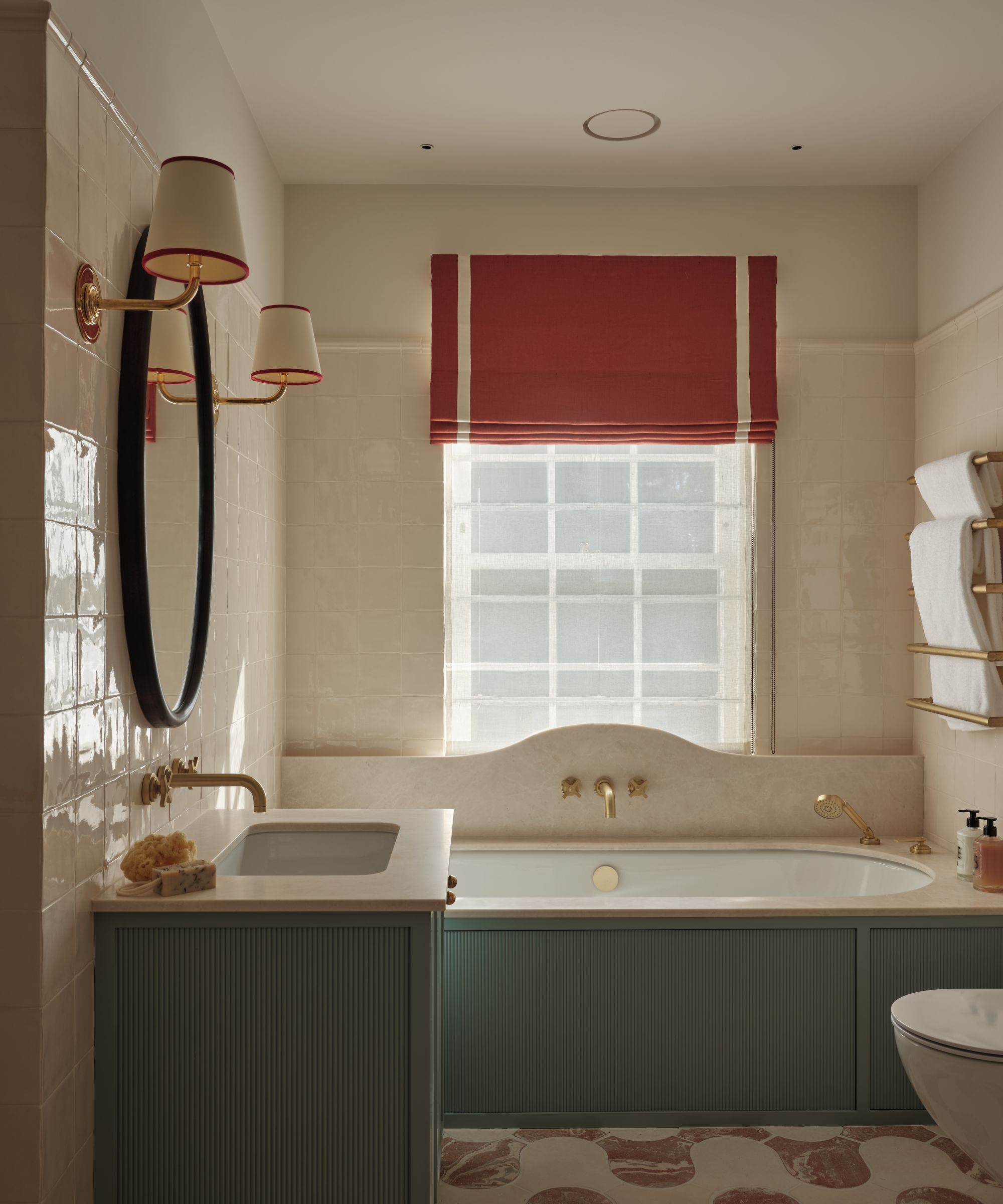 Bathroom with blue panelled bathtub and sink, marble tiled floor, cream ceramic tiled walls and oval mirror with sconces and window with red blind