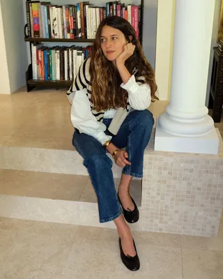 Fashion influencer Sara Ramen sits on stairs posing in a striped sweater over the shoulders, a white top, cropped straight-leg jeans, and black ballet flats.