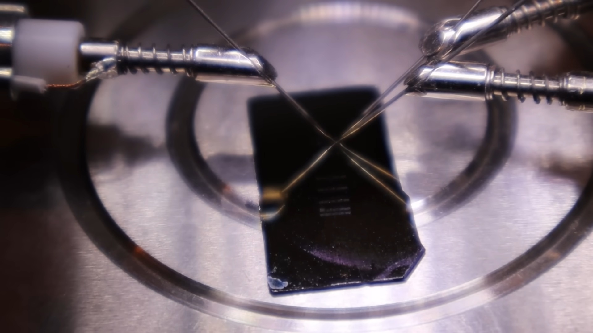 A close-up of a microchip being probed by metallic needles on a circular platform. The setting appears technical, highlighting precision and advanced technology.
