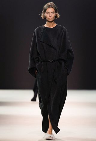 One of the best jacket coat brands, COS, is shown in an image of a model wearing a long black coat with shearling flats