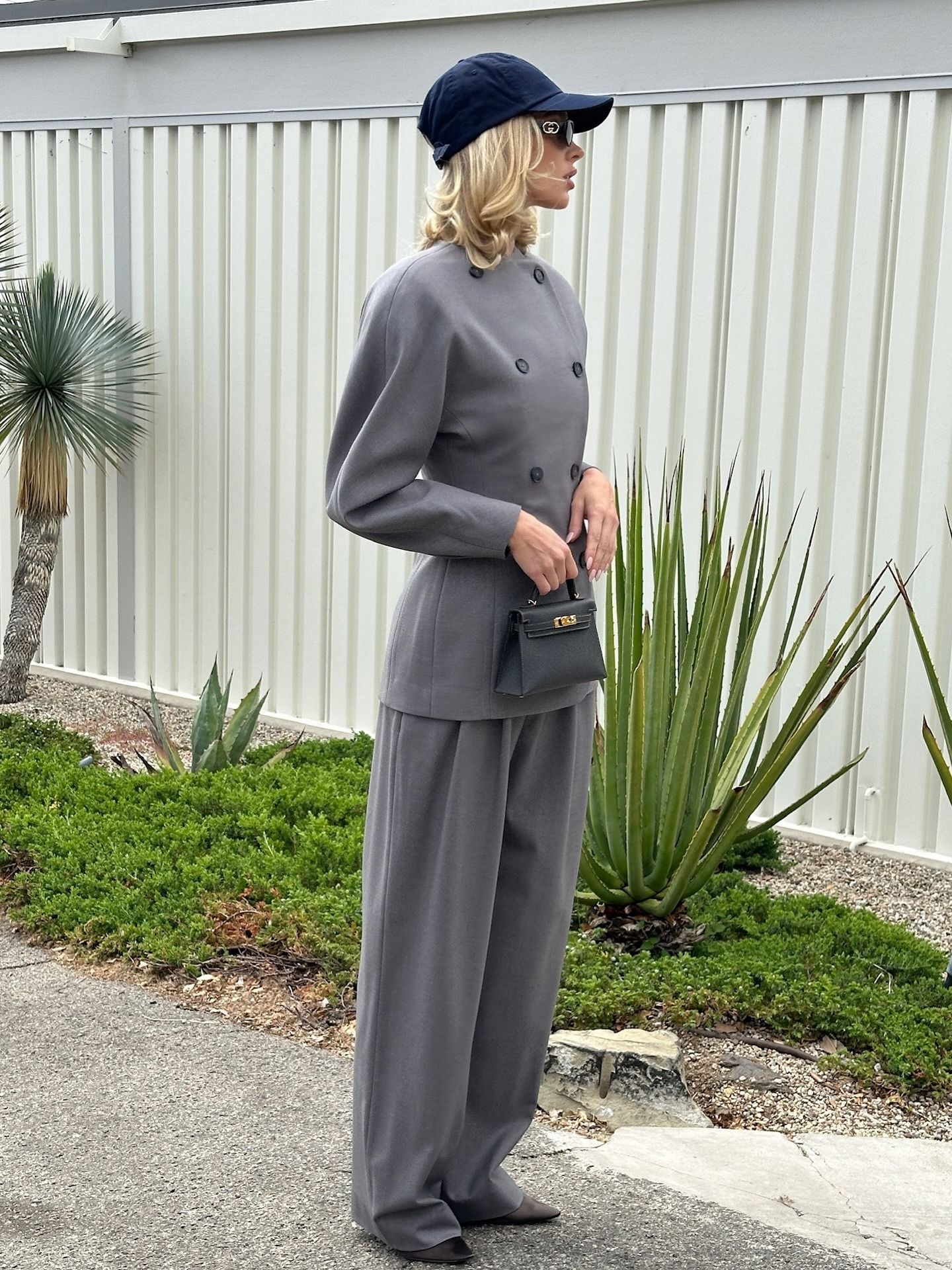 Model @hoskelsa wears a grey suit with dark brown heels and a navy cap.