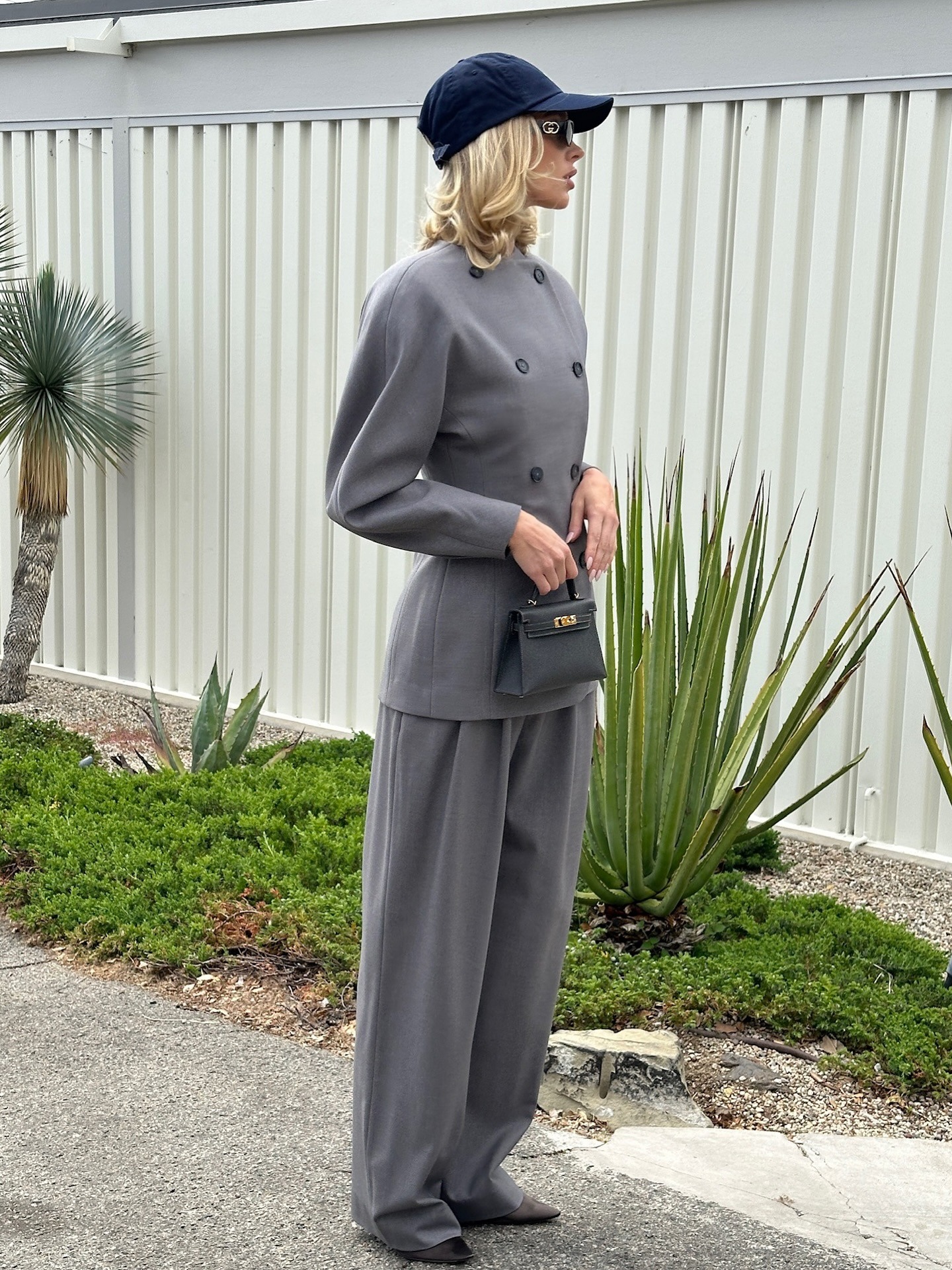 Model @hoskelsa wears a grey suit with dark brown heels and a navy cap.