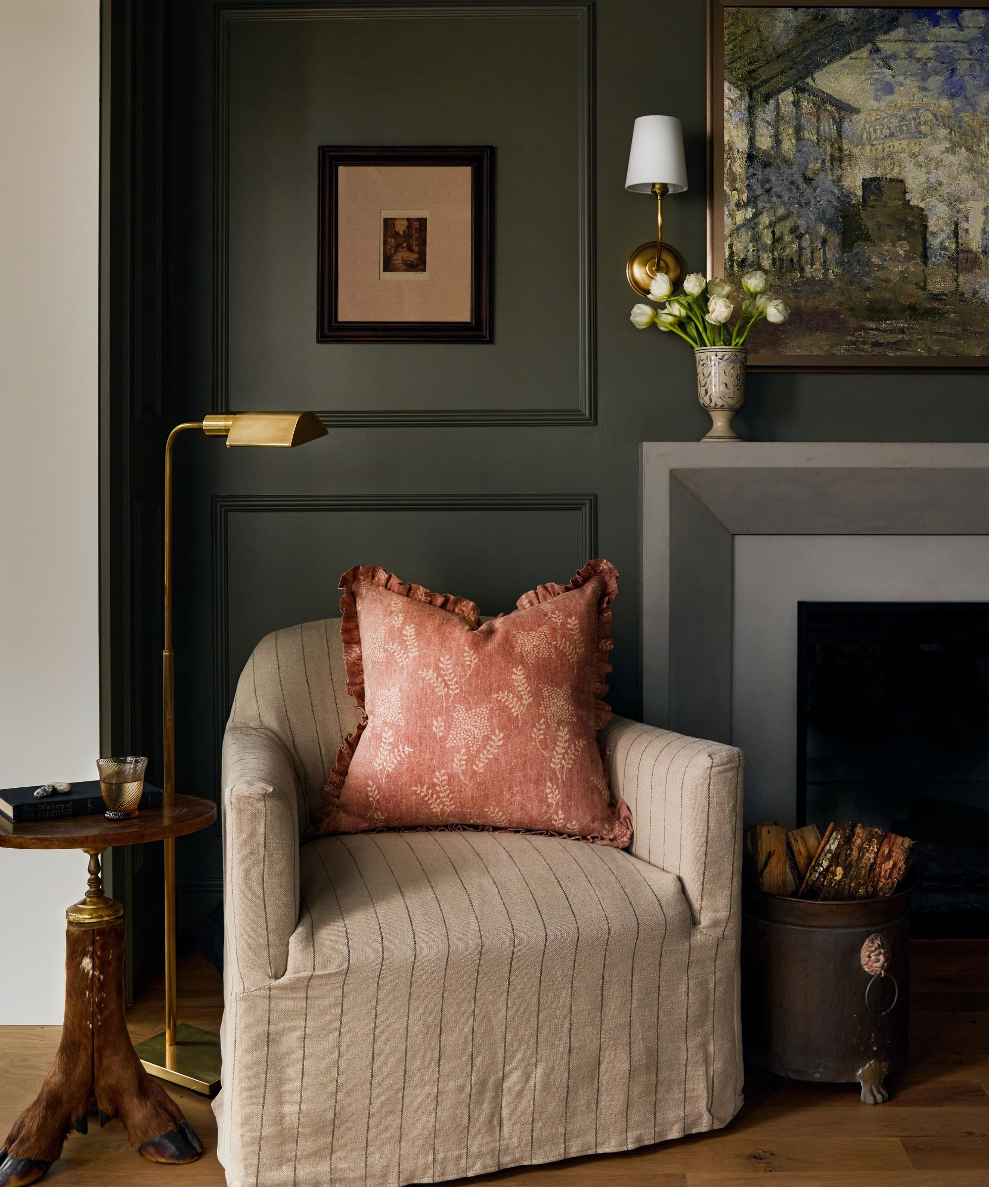 dark green living room with paneled walls, a grey fireplace with painting above, and a reading corner with a striped natural slipcovered chair with a side table and floor lamp