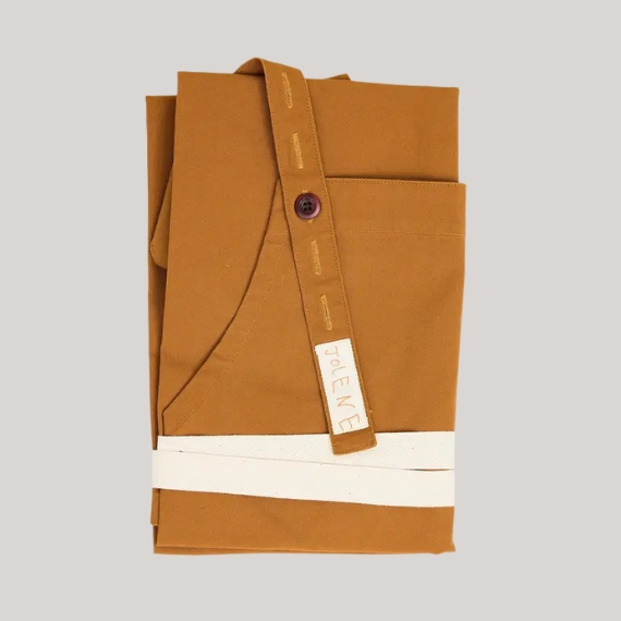 A folded apron in caramel and cream boasts the handwritten logo "JOLENE" on one of its shoulder straps while sitting against a pale grey background.