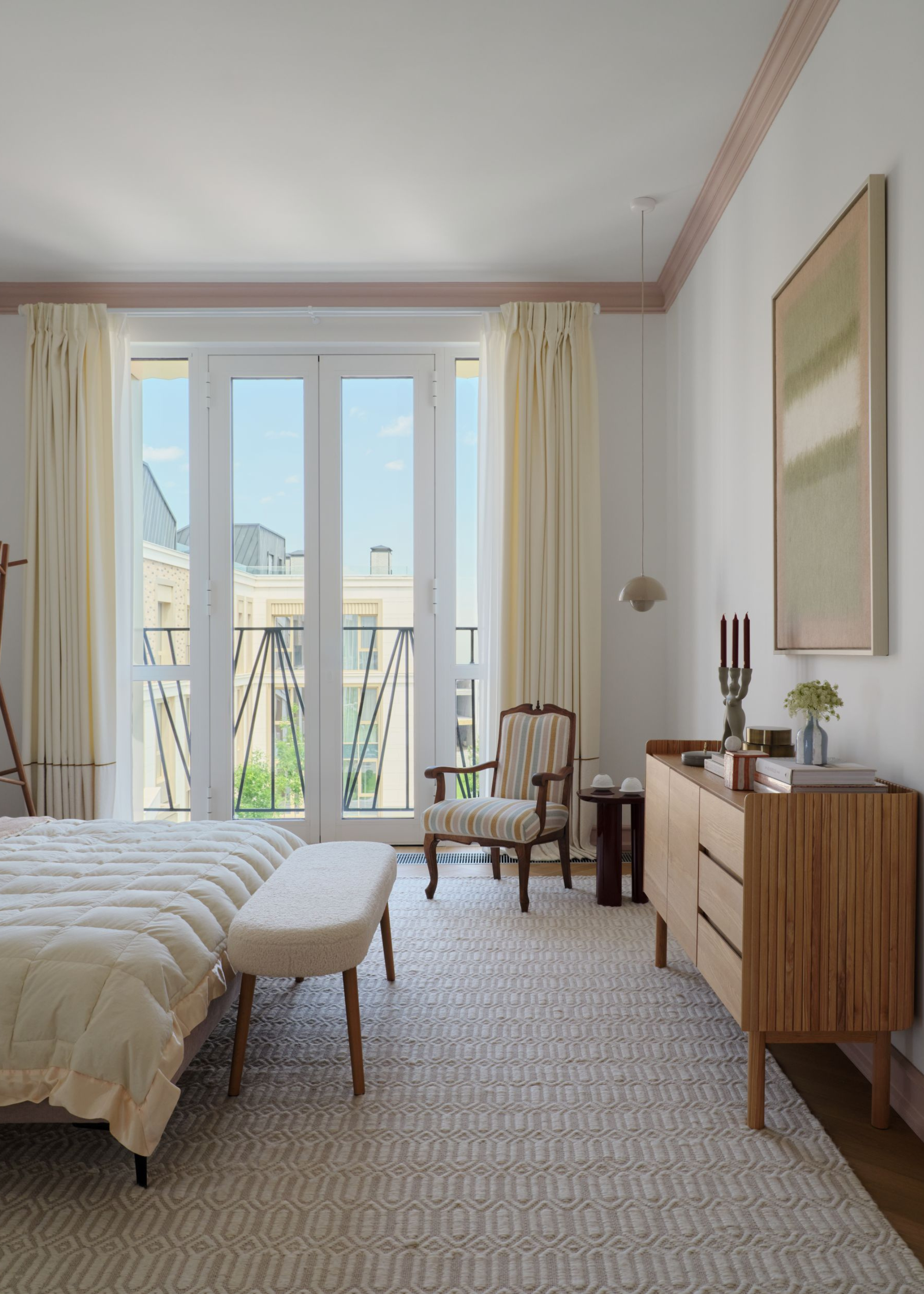 light neutral bedroom with a small white bench at the end of the bed and a wooden armchair in the corner next to the large window