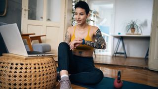 Woman sitting on an exercise mat looking at laptop, a kettlebell is on the floor next to her