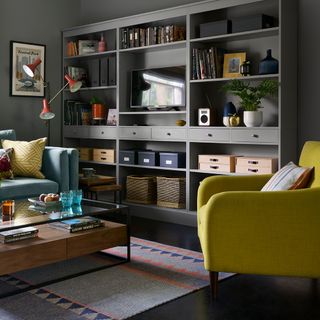 a large open living room with grey walls, a large grey wall storage unit containing an assostment of boxes, books, and items, with a blue and yellow coach in the middle of the room, and geometric pattern rug underneath a brown coffee table with black frame and glass top