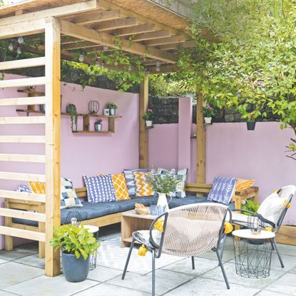 Outdoor seating area with cushions beneath wooden pergola with festoon lights next to pink garden wall