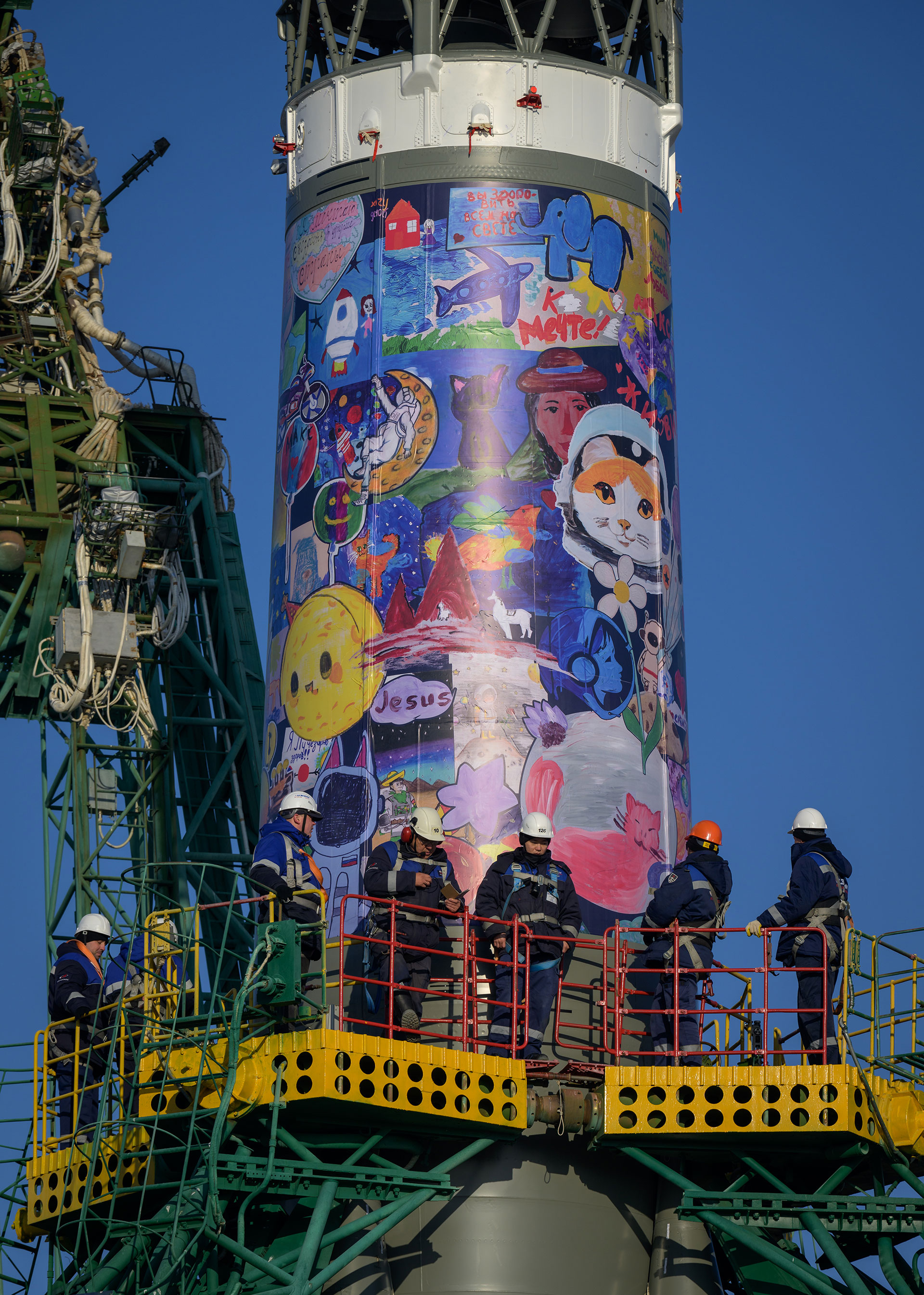 a colorful mural featuring children&#039;s drawings is seen wrapped around the body of a rocket on its launch pad