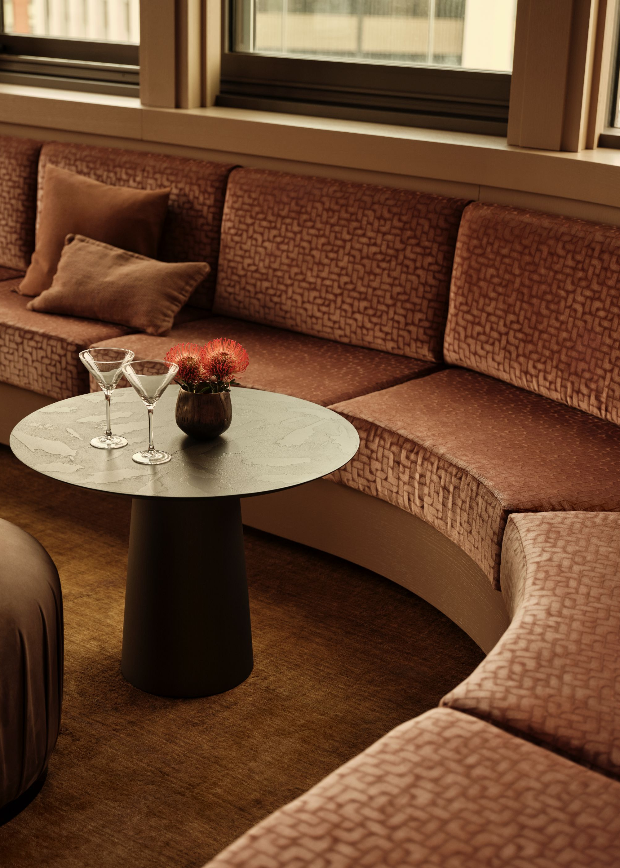 Curved banquette seating in warm, earthy tones with a round black coffee table in the corner of the curve with two empty martini glasses on top and a small vase of vibrant florals
