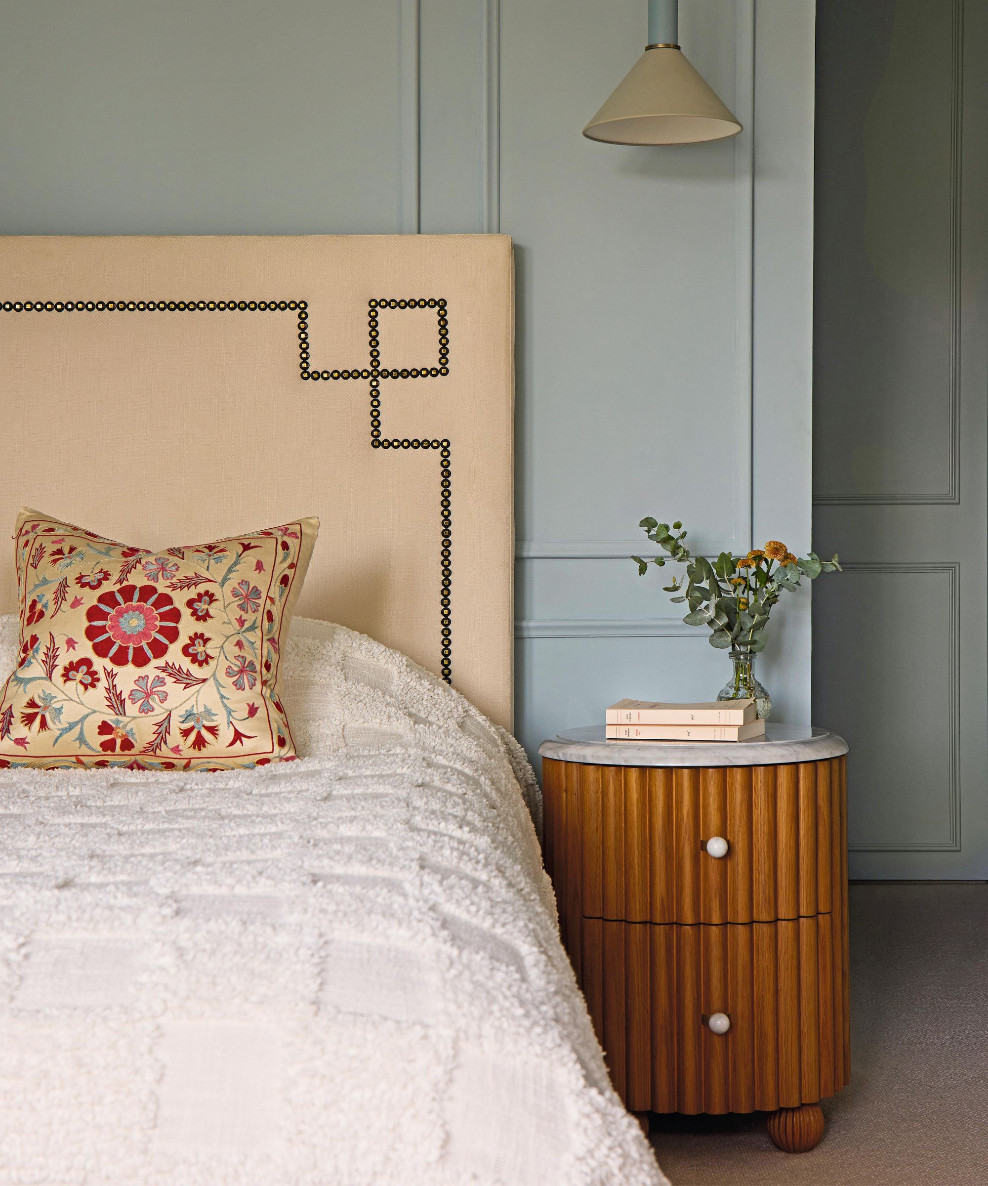 a light blue bedroom with a linen studded headboard, white textured bedding, and a fluted wood nightstand with ball feet