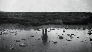 A black and white photo of an empty riverbank, with a man's figure reflected on the water