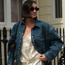 British style influencer Sarah Louise Blythe poses in London wearing black sunglasses, a denim jacket, lace-trim silk top, and lace-trim silk shorts