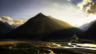 Sunlight illuminates a majestic mountain range, casting shadows over winding rivers and lush valleys under a dramatic sky