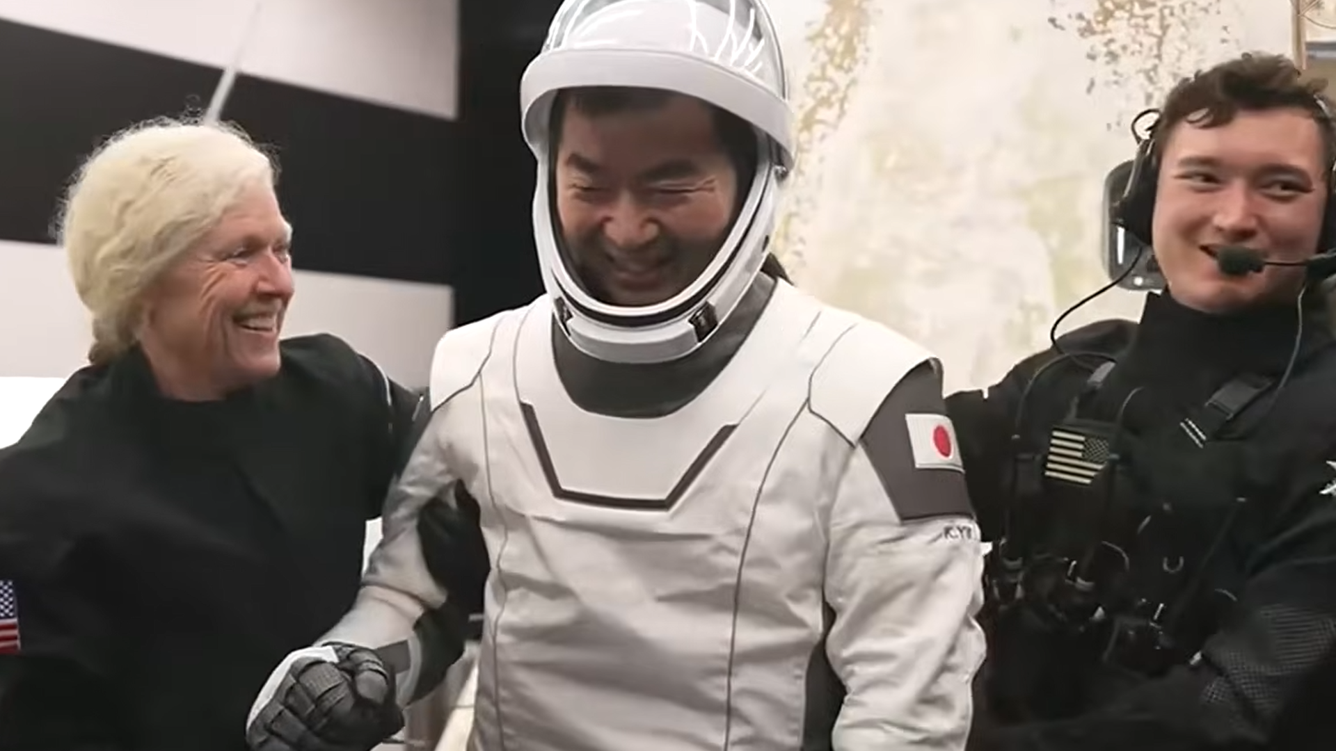 a man in a white spacesuit smiles as he is helped to his feet