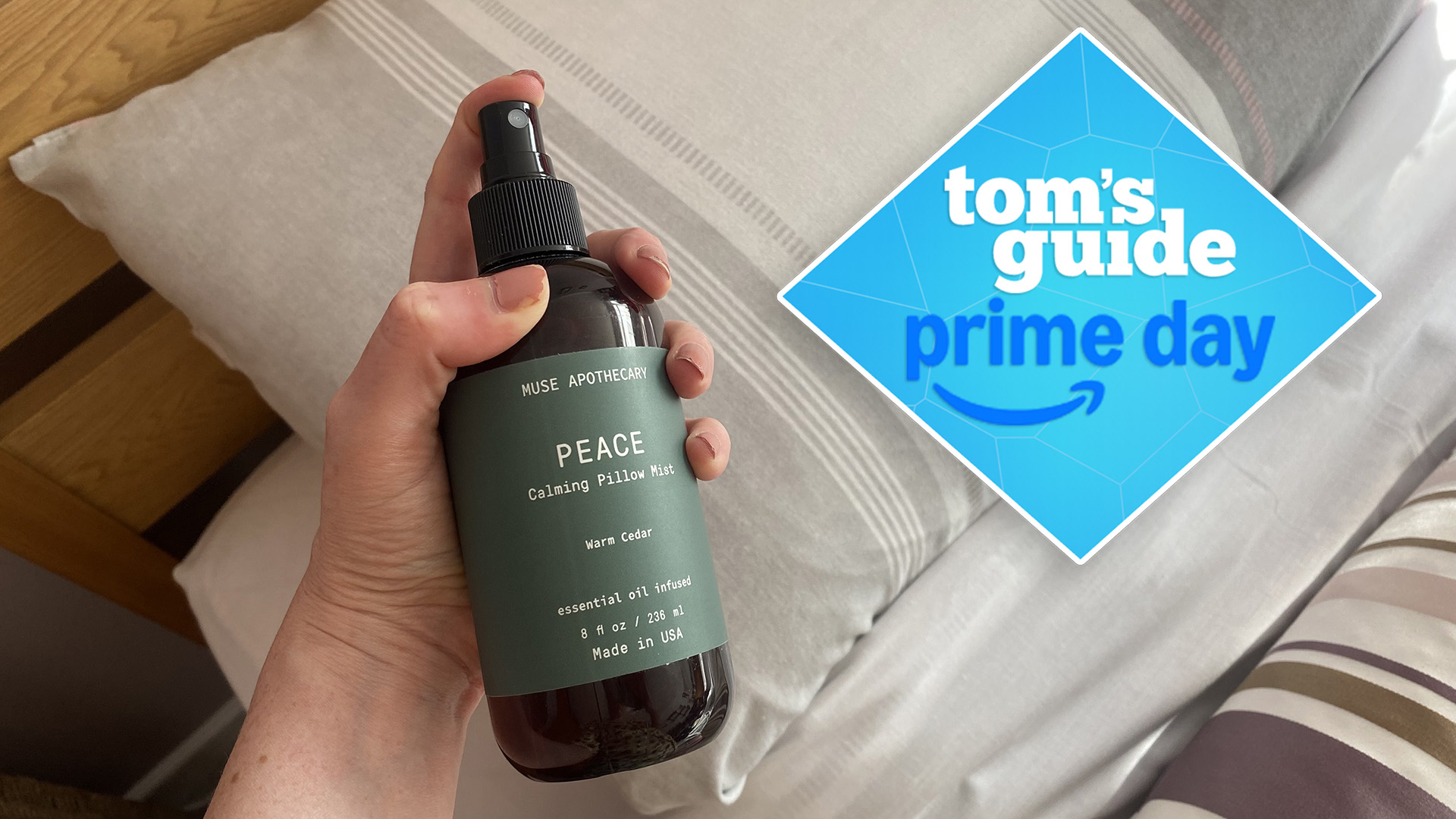 A close up of a hand holding a bottle of Muse Apothecary Peace Pillow Spray, ready to spray the soothing scent on their bedding. A Tom&#039;s Guide Prime Day deals badge in the top left corner