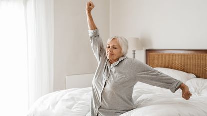 An older woman sitting on the side of her bed stretches after waking up.