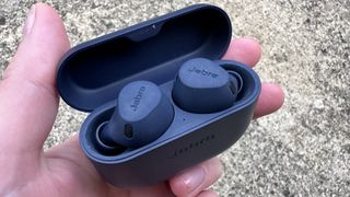 Someone holding the Jabra Elite 8 Active in their charging case in dark blue above some concrete