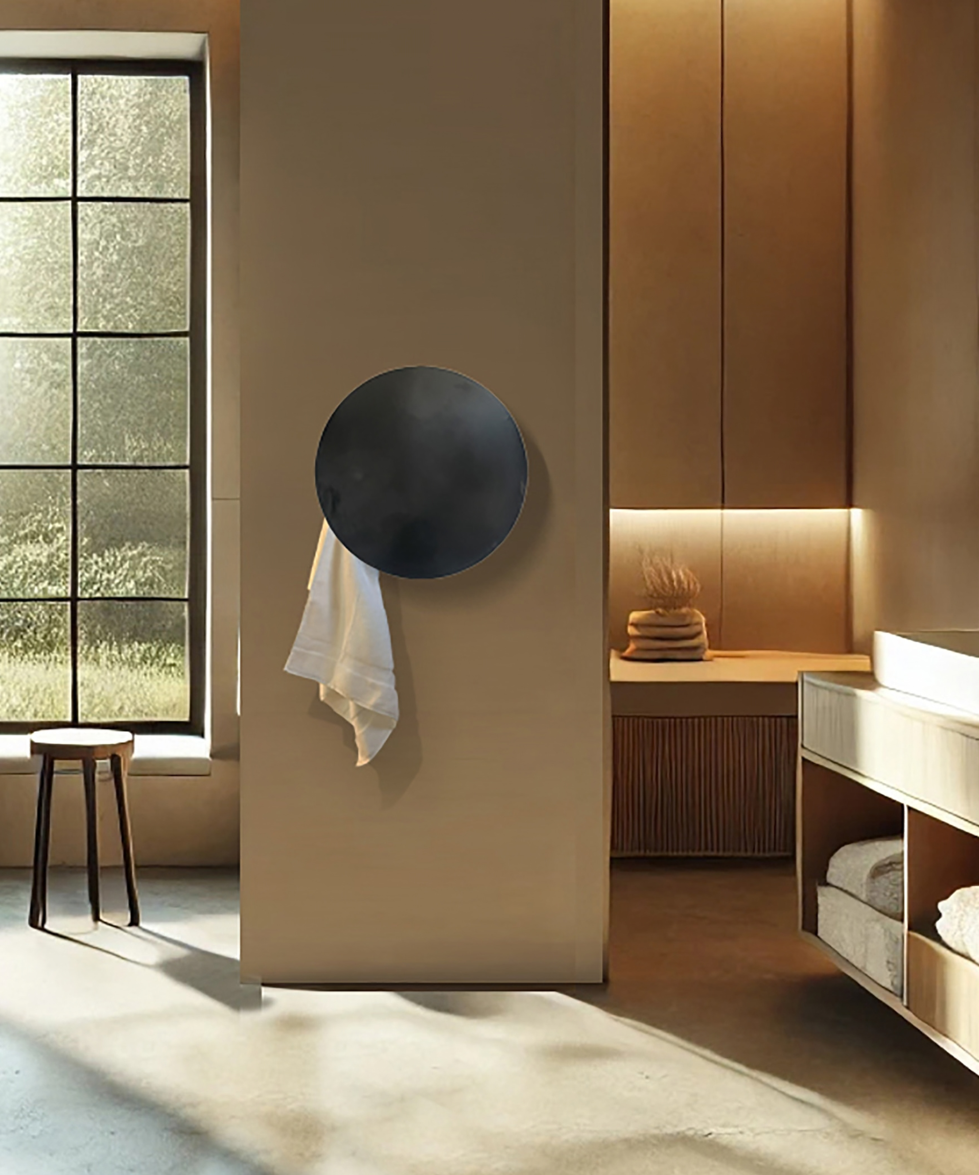 A sculptural matte black circular heated towel radiator mounted on a smooth taupe wall, with a single white towel draped from its lower edge. Behind, a warm minimal bathroom features oak cabinetry with under-cabinet lighting, a wooden stool, and a large steel-framed window overlooking greenery.