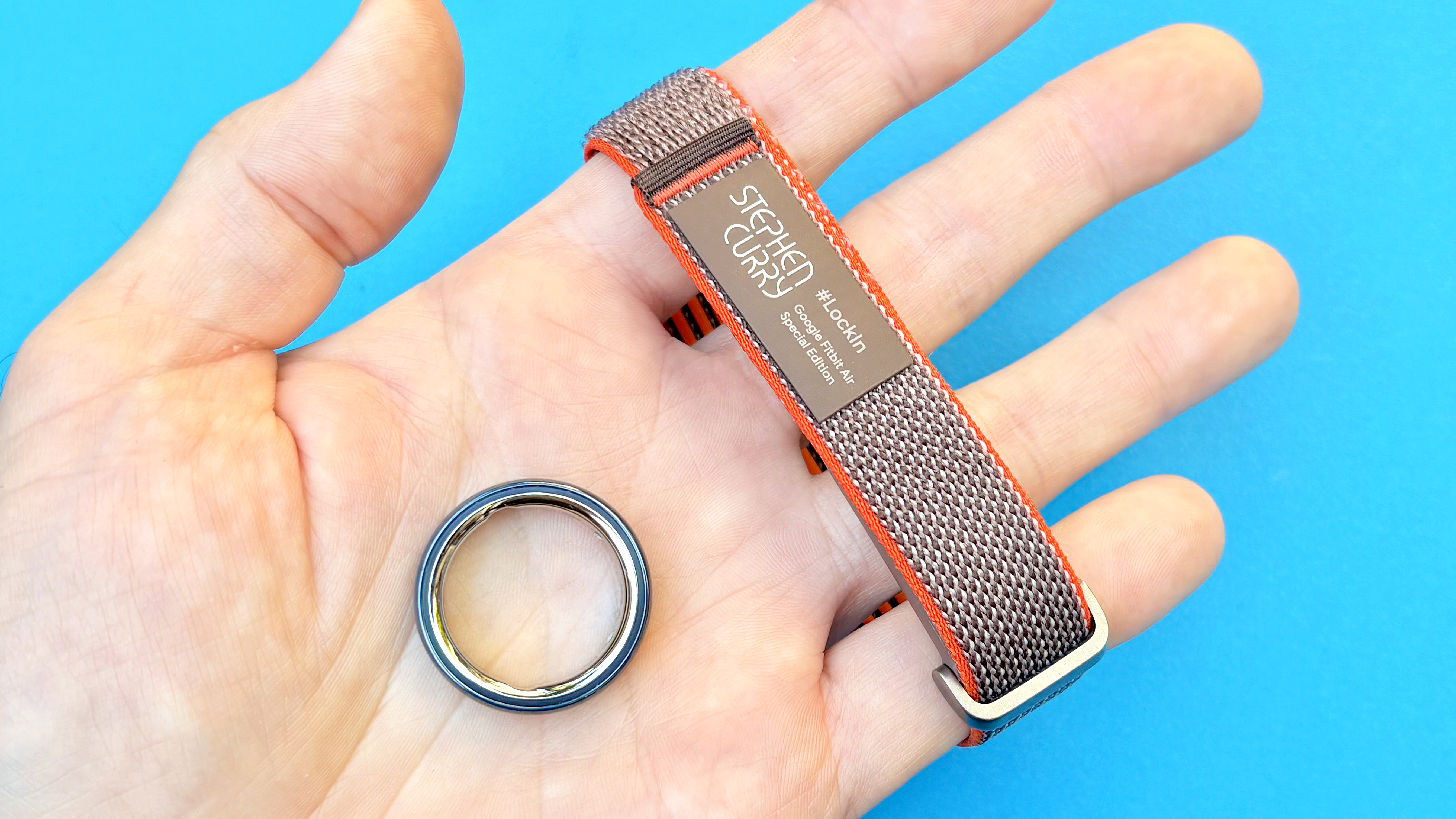 Close-up of the Fitbit Air and Oura Ring 4 Ceramic in the writer's hand against a blue background