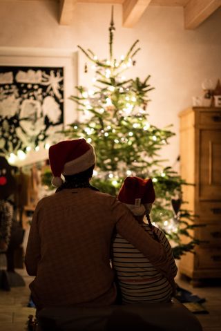 The silhouettes of a parent and young child both wearing Santa hats. They are out of focus, with a christmas tree lit in gold lights in focus in the background