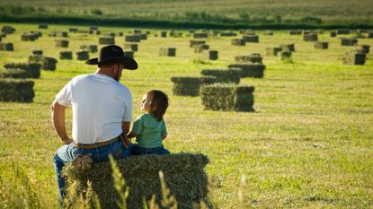 A farmer and his little girl sit on a hay bale overlooking a huge field.