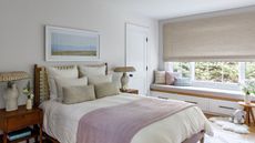 Small bedroom with bright window and natural accents. There is a wooden bed and nightstand, with a bench seat under the window