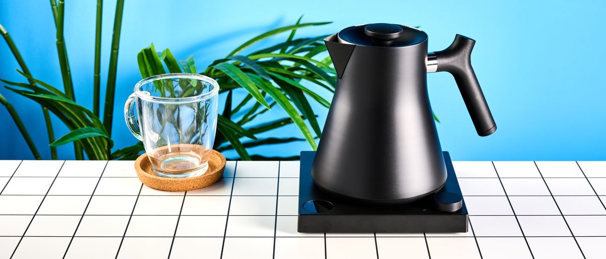 the fellow Corvo EKG kettle in matte black photographed against a blue background with a glass mug and black and white table 