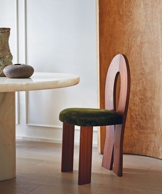 Close up of sculptural dark wood and green dining chair, curved wood screen in background, curved marble dining table with two decorative vases