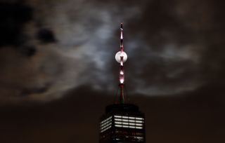 A tall building in the foreground with the moon shining behind it in the background. The skies are dark and covered with patchy clouds.