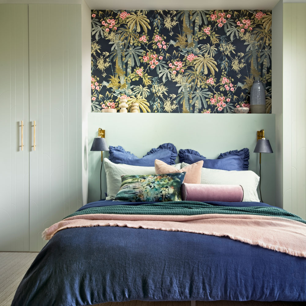 Small bedroom with built in cupboards, double bed, blue duvet, colourful cushions and floral wall painting