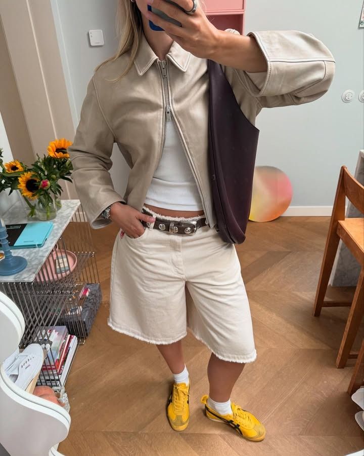 A woman standing wearing white baggy jean shorts, a studded belt, a leather jacket, and yellow sneakers.