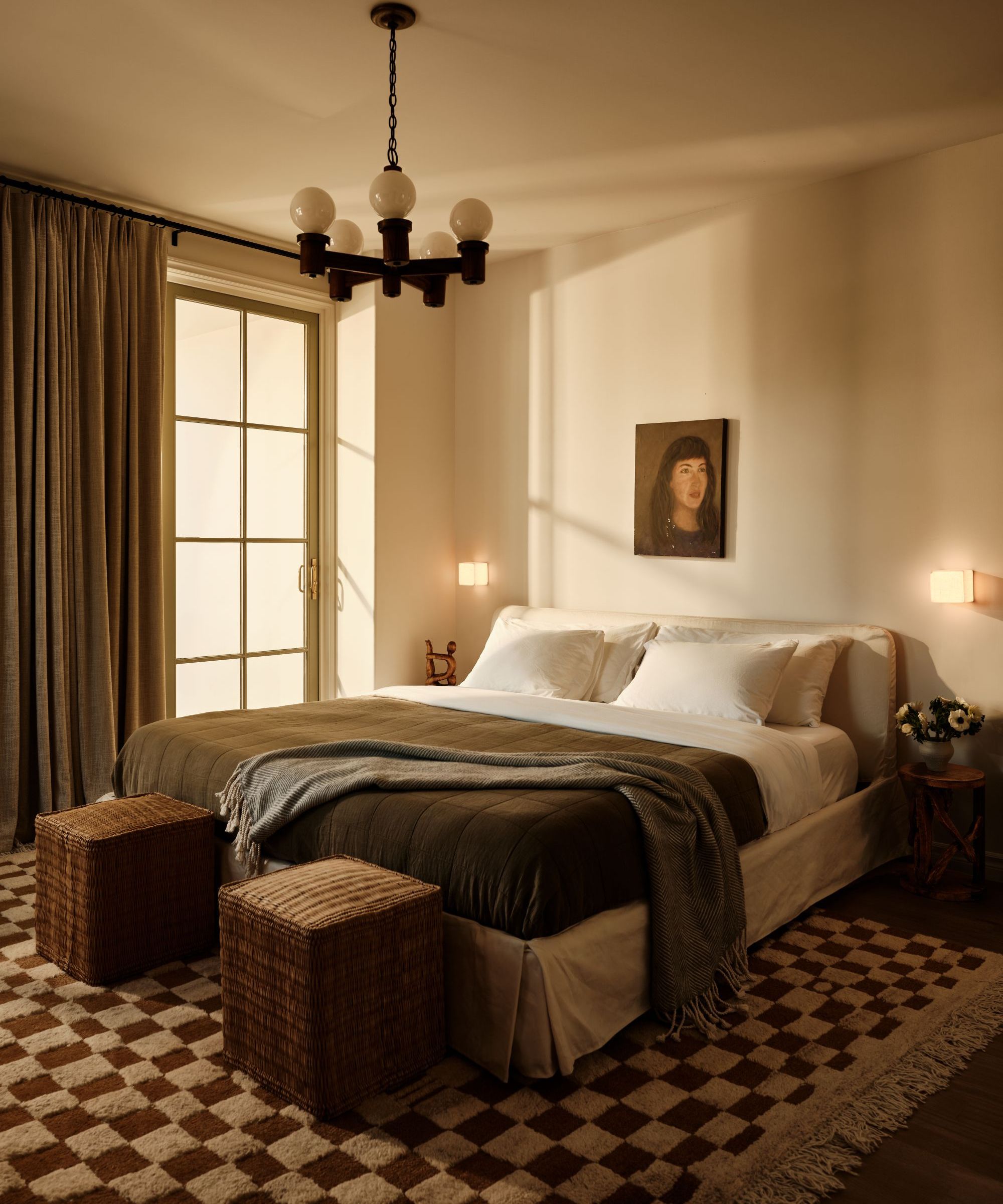 a bedroom with a checker printed rug, two ottomans at the foot of the bed, a dark green quilt, and white sheets on the bed, a portrait hanging on the wall above, and curtains covering the sliding glass door