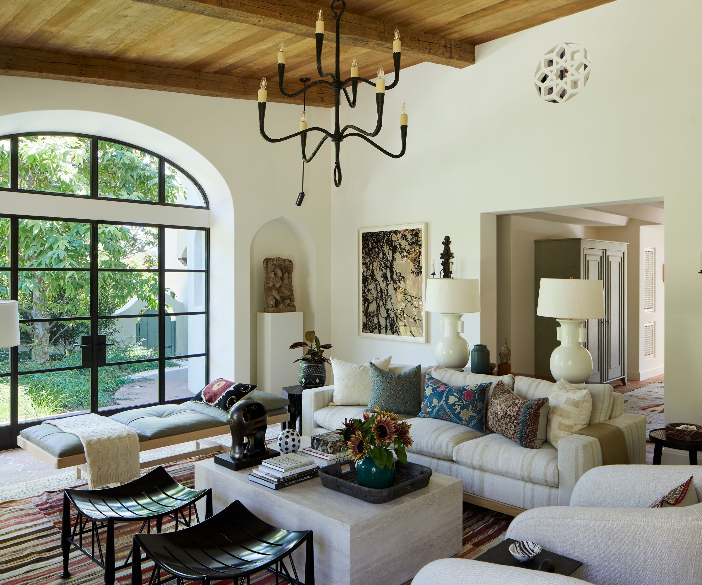 Transitional living room with arched glass doors, wrought-iron chandelier, white walls, beige striped sofa, curved cream armchair, stone ottoman with decor, a mid-century daybed, and two leather black stools on top of a striped rug