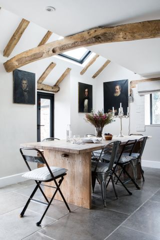 rustic wooden dining table with black chairs in room with white walls with beams and old master paintings on wallsand grey floor