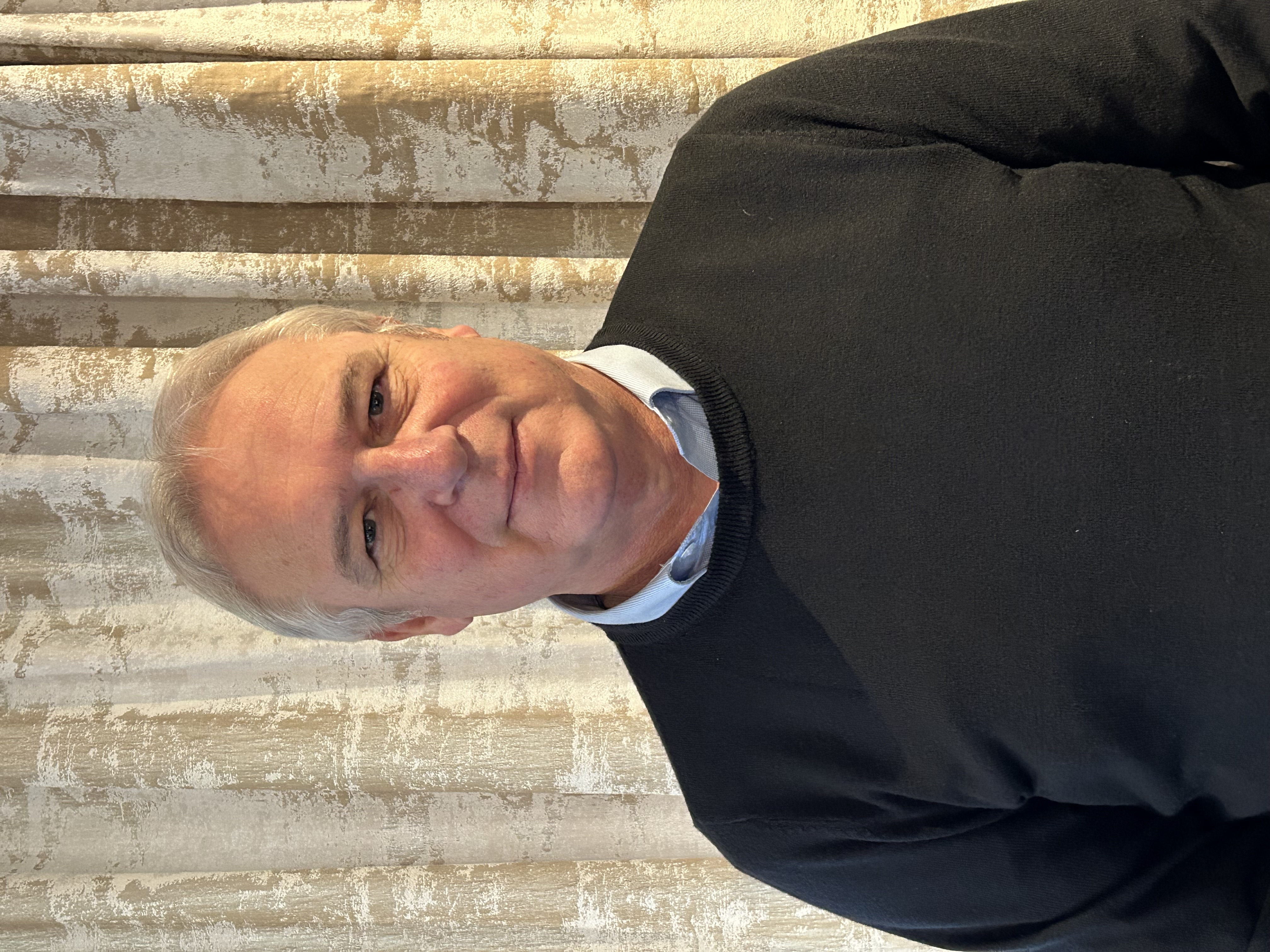 A headshot of Richard Bradley, standing in front of cream patterned curtains looking at the camera with gray hair and wearing a black jumper with a blue shirt underneath