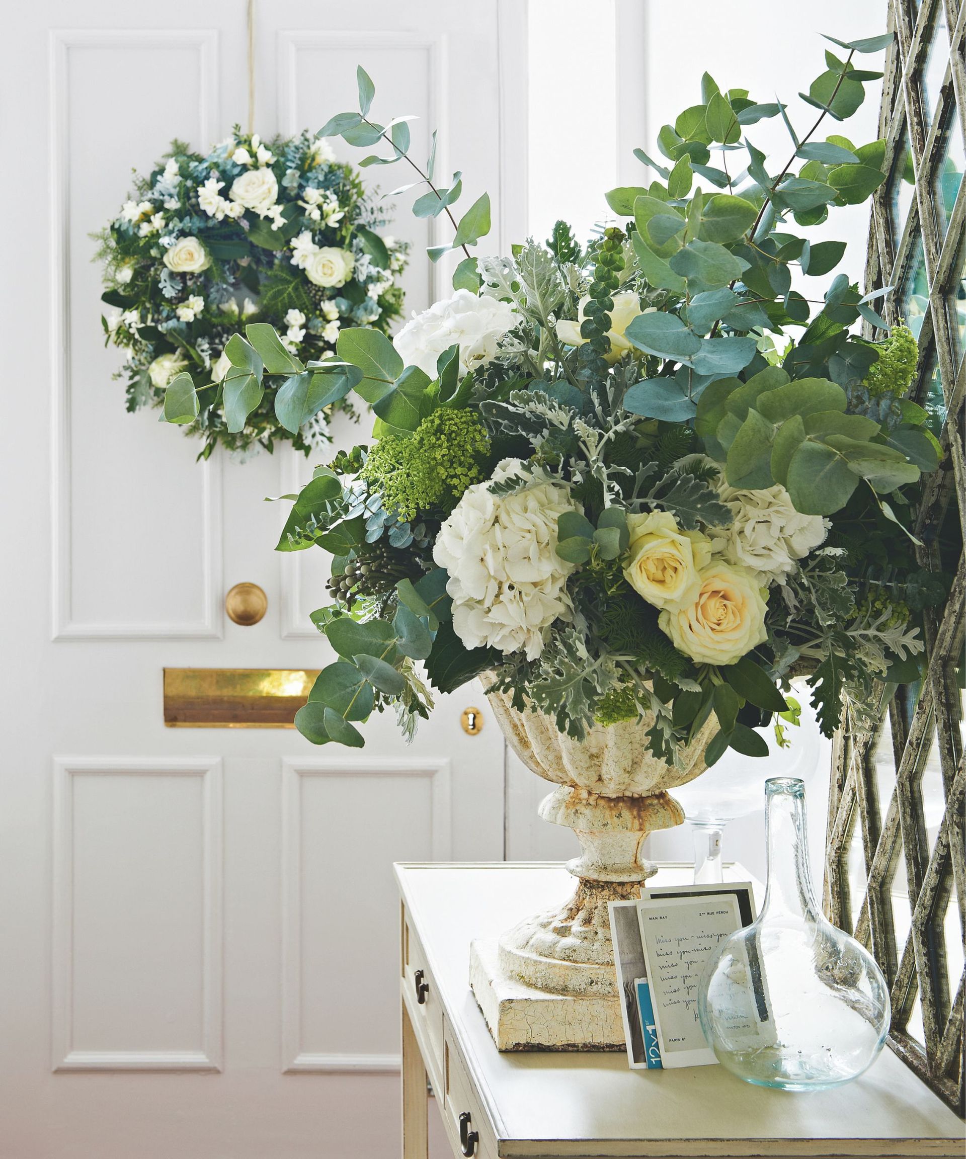 urn of white and green flowers and foliage on a hallway table with a white doorway in shot and a white and green wreath hanging on it
