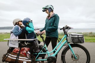 The best electric cargo bikes allow you to travel as a whole family. This image shows two children on the back of an electric cargo bike while a woman talks to them off bike