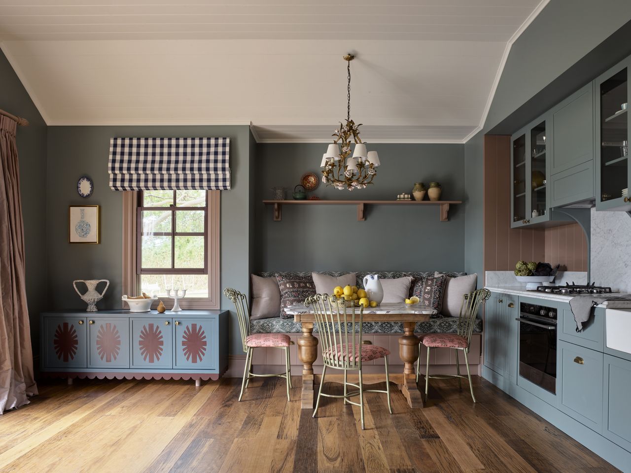 colorful kitchen with blue cabinetry, pink backsplash, and built-in dining nook with shelf overhead, and pendant light, timber floorboards, and pink window frames, with a hand-painted console styled with vases and a checkered roman blind