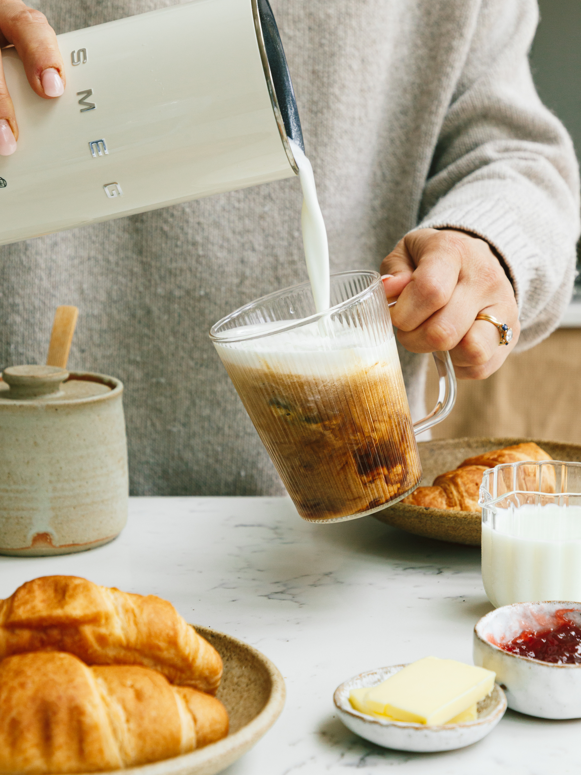Image of a hand over a marble countertop pouring milk from a white Smeg milk frother into a fluted glass that has an iced coffee in it.