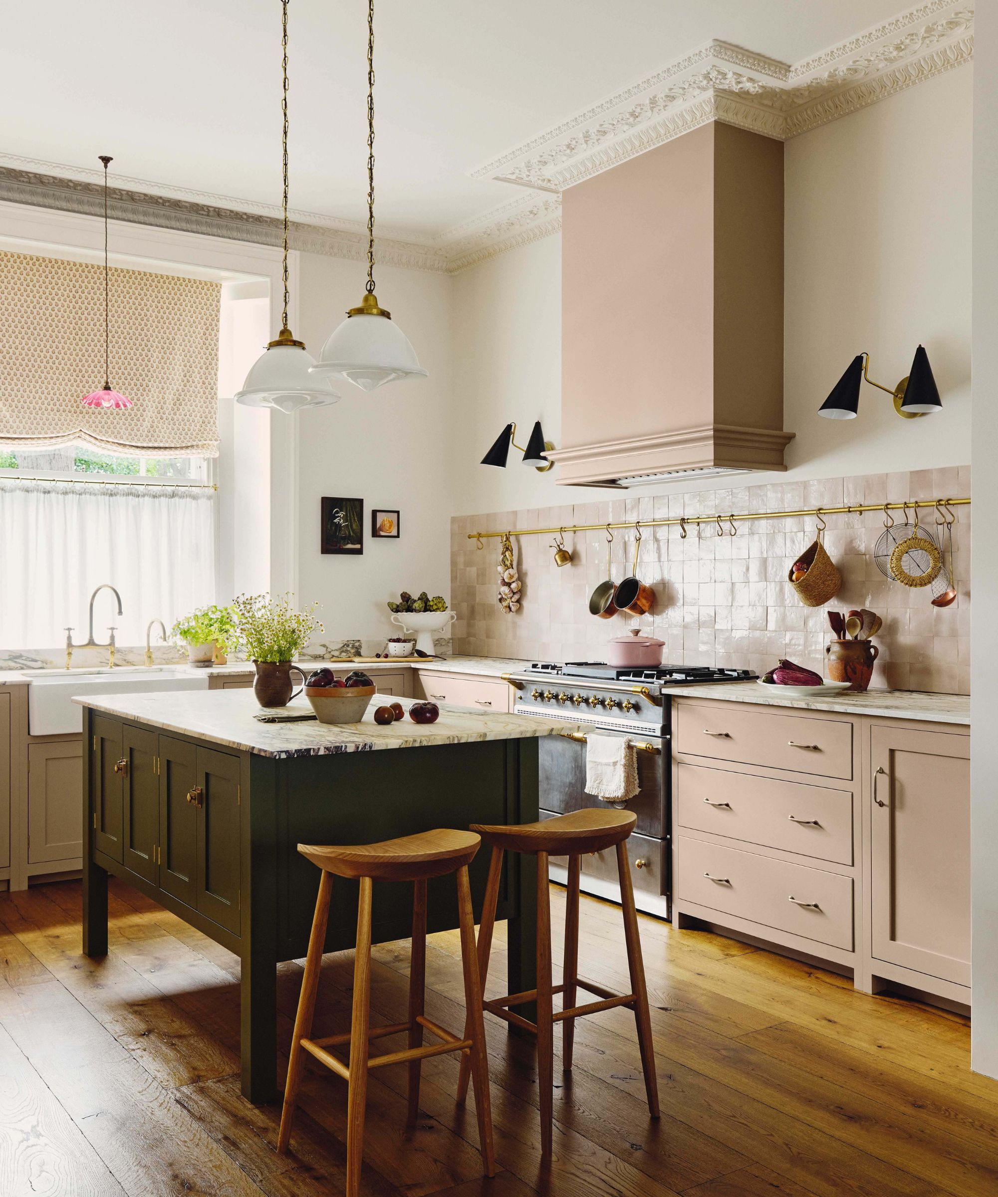 A spacious kitchen with pink lower cabinets, a dark green central island with wooden barstools, and a large range cooker under a matching mauve vent hood